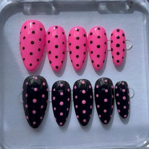 Puede incluir: Un conjunto de uñas postizas en dos diseños. Un juego es rosa brillante con lunares negros, y el otro es negro con lunares rosas. Las uñas tienen forma ovalada y están dispuestas en filas.