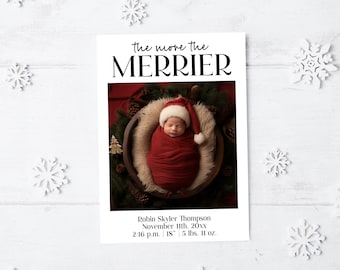 May include: A Christmas-themed birth announcement card with the text "the more the MERRIER." The card features a newborn baby wrapped in a red blanket and wearing a Santa hat. The baby is in a wooden bowl surrounded by greenery. The card includes the baby's name, birth date, time, height (18 inches), and weight (5 lbs. 11 oz.).