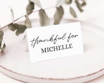 Puede incluir: Una tarjeta de mesa blanca con las palabras "thankful for MICHELLE" en escritura negra y fuente sans-serif. La tarjeta está sobre un plato blanco con una ramita de vegetación en el fondo.
