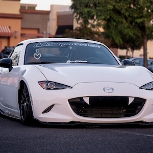 May include: White Mazda MX-5 with black accents and a custom front bumper with fangs. The car has a heart sticker on the windscreen and a "#damped" sticker on the top of the windscreen.