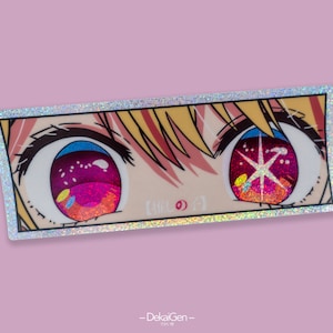 May include: A close-up of a holographic sticker featuring a pair of anime eyes with pink and red irises. The right eye has a white star in the center. The sticker has a silver border and a glittery finish.