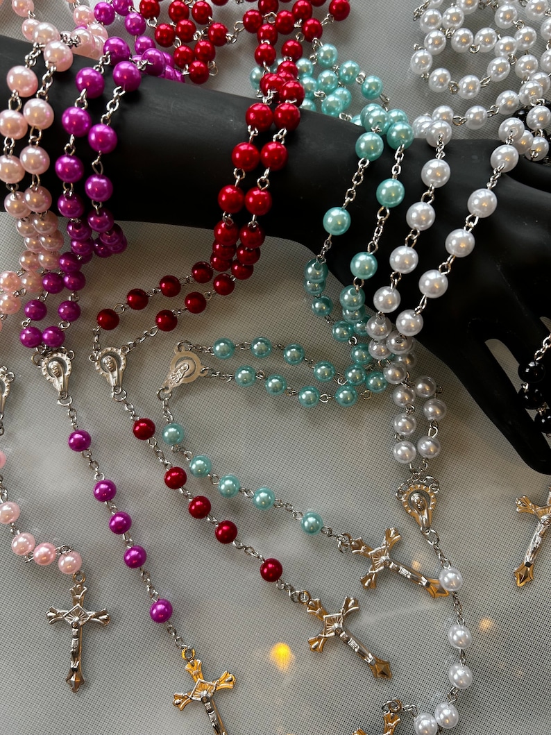 May include: A set of five pearl rosaries with silver crosses and charms. The rosaries are in pink, purple, red, blue, and white.