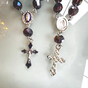 Op de afbeelding: Twee rozenkransen met donkerpaarse facetkralen en zilverkleurige metalen accenten. Elke rozenkrans heeft een kruisvormige hanger en een centraal medaillon met religieuze afbeeldingen. De kruisen hebben een donkerblauwe emaille afwerking.