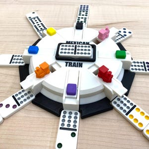 May include: A white Mexican Train dominoes game set with colorful train markers and dominoes arranged around a central hub. The hub has the words "Mexican Train" printed on it. The game is set on a light brown wooden surface.