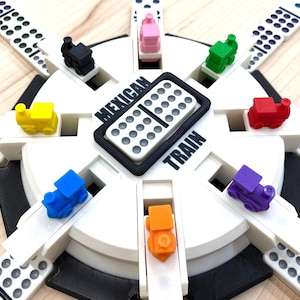 May include: A Mexican Train dominoes game set. The game features a white circular base with slots for dominoes and colorful toy train markers. The center displays the words "Mexican Train".