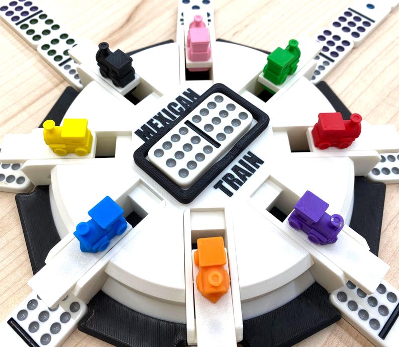 May include: A Mexican Train dominoes game set. The game features a white circular base with slots for dominoes and colorful toy train markers. The center displays the words "Mexican Train".