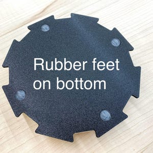 May include: Black, geometric-shaped object with a textured surface. The text "Rubber feet on bottom" is printed in white. Four small, gray rubber feet are visible on the bottom of the object.