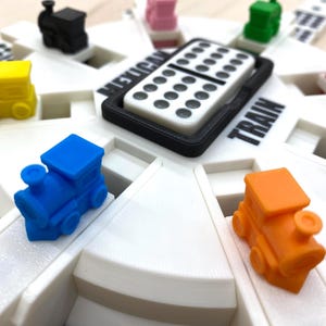 May include: A white Mexican Train dominoes game board with colorful miniature train markers. The board has the words "Mexican Train" and "Train" printed on it. A black tray holds a domino.