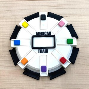 May include: A white and black Mexican Train dominoes game board with the words "MEXICAN TRAIN" printed on it. The board has a rectangular black frame in the center and colored pegs in yellow, blue, pink, green, red, orange, and purple.