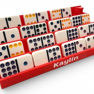 May include: A red domino rack holding white dominoes with colored dots. The dominoes are arranged in rows, displaying various dot combinations. The word "Kaylin" is printed on the side of the rack.
