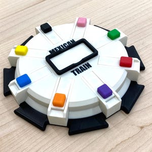 May include: A white and black Mexican Train dominoes game with colorful markers. The game board has a circular design with the words "Mexican Train" printed on it. The game includes a black frame in the center and colorful markers.