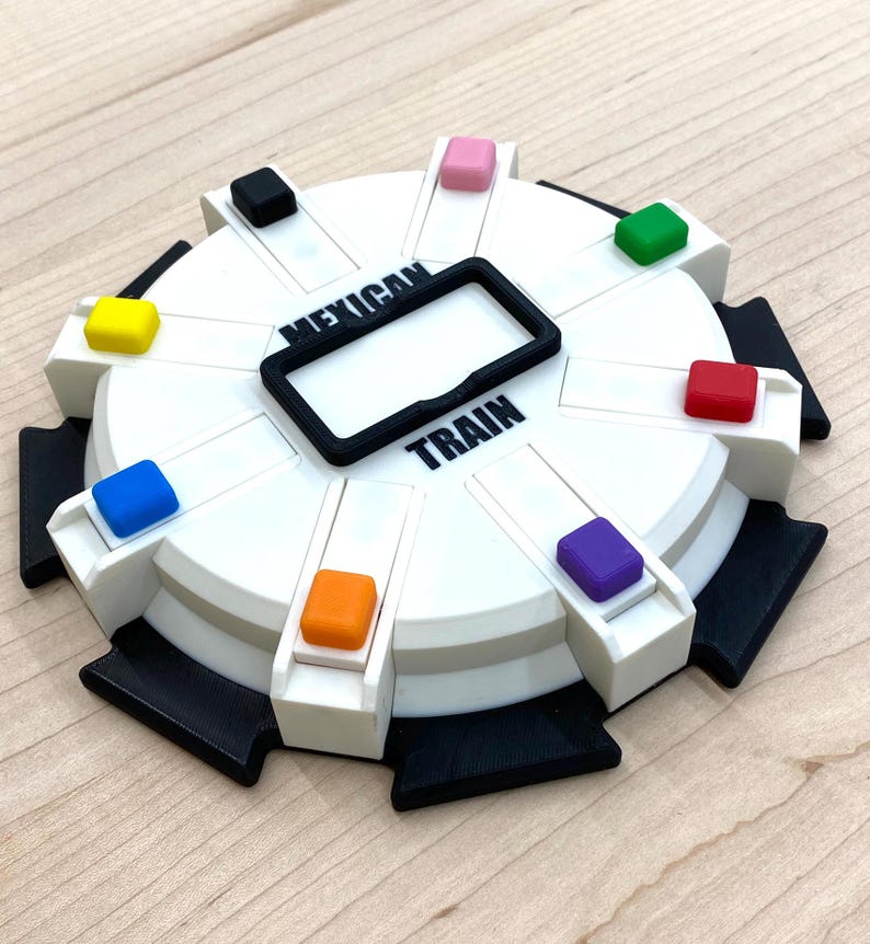 May include: A white and black Mexican Train dominoes game with colorful markers. The game board has a circular design with the words "Mexican Train" printed on it. The game includes a black frame in the center and colorful markers.
