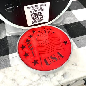May include: A red, circular lid with a patriotic design featuring the word "USA", stars, a rocket, and a flag. The lid is part of a slush machine. The text "FOR THE BEST SLUSH, SCAN BELOW TO GET STARTED" is visible.