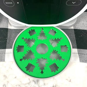May include: A green, circular insert with Christmas tree and square cutouts. The insert sits beneath a black and white appliance with the word "RINSE" visible. The background is a black and white checkered pattern.