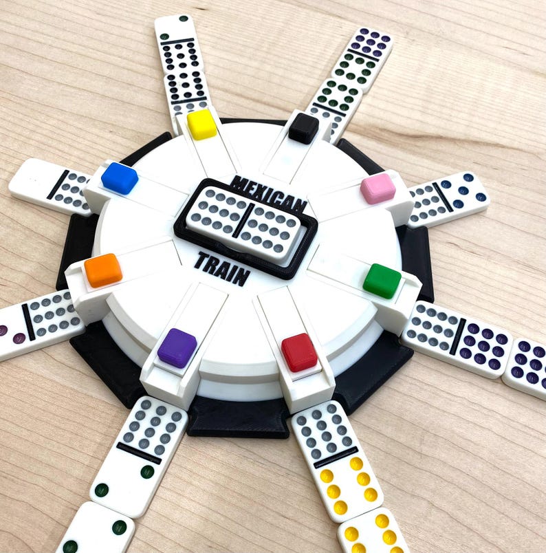 May include: A white and black Mexican Train dominoes game set. The set includes a central hub with colored markers and dominoes arranged around it. The text "MEXICAN TRAIN" is printed on the hub.