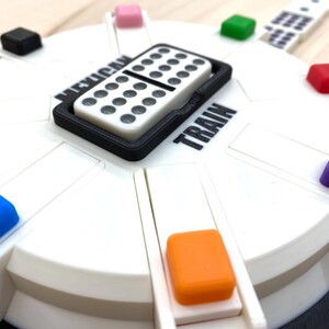 May include: A white and black Mexican Train dominoes game set. The game board is white with colorful markers and the words "Mexican Train" and "Train" printed on it. A domino is in a black tray.