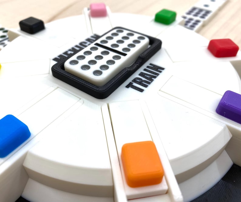May include: A white and black Mexican Train dominoes game set. The game board is white with colorful markers and the words "Mexican Train" and "Train" printed on it. A domino is in a black tray.