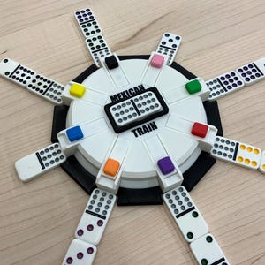 May include: A Mexican Train dominoes game set. The game features a white and black circular hub with colored markers and the words "Mexican Train". White dominoes with black and colored dots are arranged around the hub.