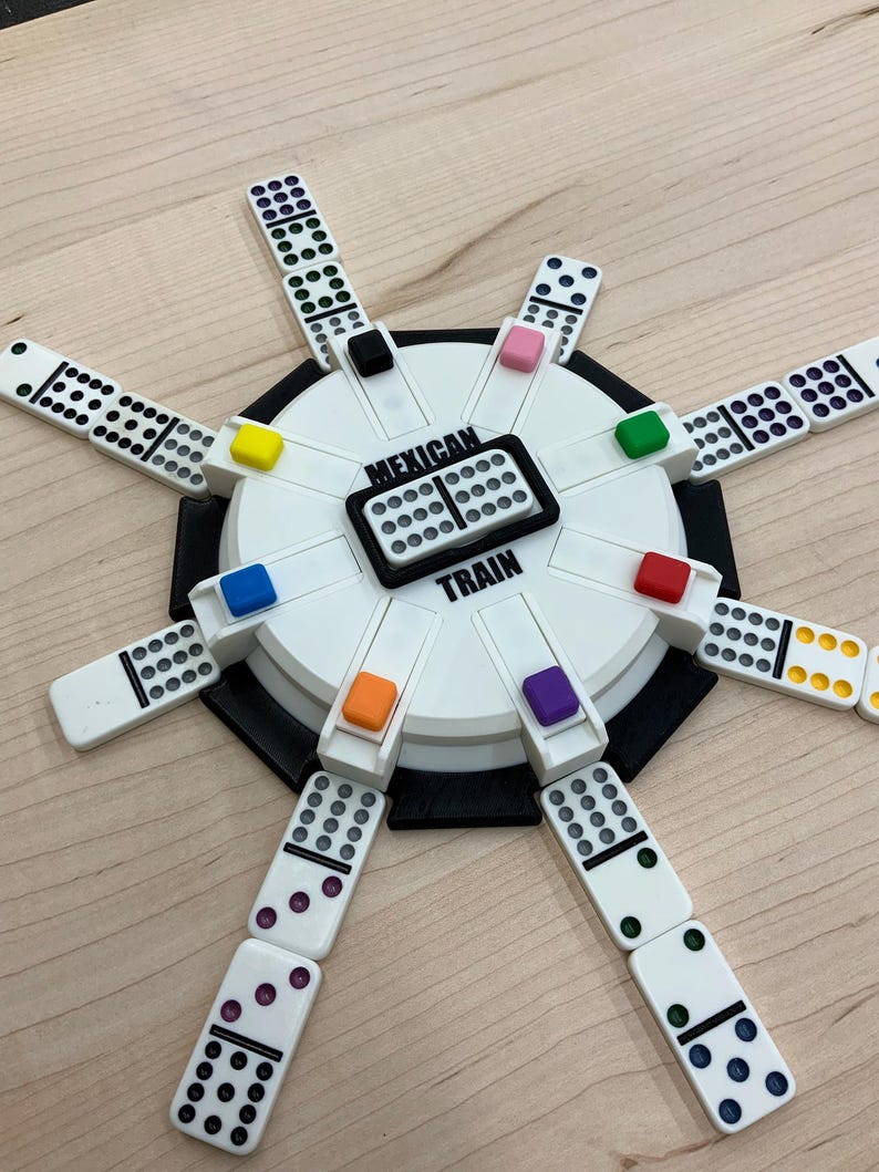 May include: A Mexican Train dominoes game set. The game features a white and black circular hub with colored markers and the words "Mexican Train". White dominoes with black and colored dots are arranged around the hub.