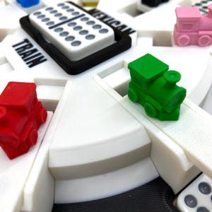 May include: Close-up of a Mexican Train dominoes game. The game features a white circular board with the words "Mexican Train" and "Train" printed on it. Colorful miniature train pieces in red, green, and pink are placed on the board, along with dominoes.