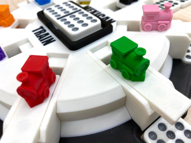 May include: Close-up of a Mexican Train dominoes game. The game features a white circular board with the words "Mexican Train" and "Train" printed on it. Colorful miniature train pieces in red, green, and pink are placed on the board, along with dominoes.