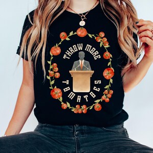 以下が含まれることがあります： トマトと「THROW MORE TOMATOES」の文字が入った円形デザインの黒いTシャツ。デザインには演台に立つ人のイラストが含まれています。トマトは赤色で、文字は白色です。
