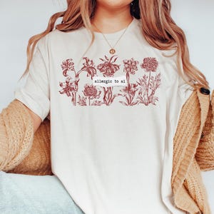 Op de afbeelding: Een crèmekleurig t-shirt met een donkerrode botanische illustratie van diverse bloemen in een horizontale rij. Gecentreerd in het bloemendesign bevindt zich een wit rechthoekig grafisch element met de zwarte tekst "allergic to ai".