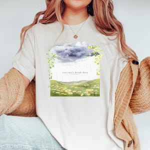 以下が含まれることがあります： クリーム色のTシャツで、黄色いデイジーと緑のツルが描かれた緑の草原の上に雨雲がプリントされています。グラフィックの中央には「you can't drink data」という文字が印刷されています。ベージュのカーディガンを羽織っています。