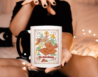 May include: White ceramic mug with a black handle and rim. The mug features a tarot card design with a hand holding cards, flowers, and the text "The Avid Reader". The design is in shades of brown, orange, and green.