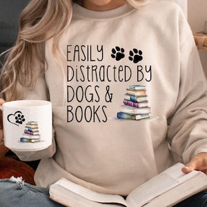 May include: A woman wearing a beige sweatshirt with the text "Easily Distracted by Dogs & Books" and a stack of books and paw prints. She is holding a white mug with a paw print and heart design. She is sitting on a floor with a book in her lap.
