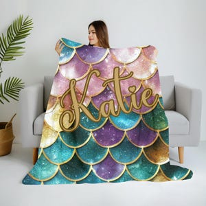 Puede incluir: Una manta de tiro con estampado de escamas de sirena en color verde azulado, morado y dorado con el nombre "Katie" escrito en letras doradas.