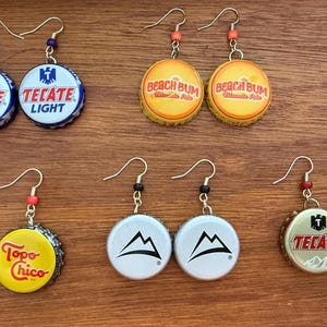 May include: Handmade earrings featuring repurposed bottle caps. Designs include "Tecate Light", "Beach Bum", "Topo Chico", and a mountain logo. Each pair has a unique hook and bead detail, offering a fun, eco-friendly accessory.