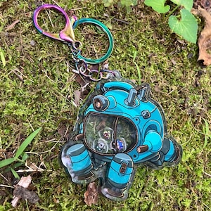 May include: A clear acrylic keychain featuring a blue robotic character with a transparent dome window. Inside the dome are three small, colorful character icons. The keychain is attached to a metallic, iridescent rainbow-colored lobster clasp.