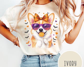 May include: Ivory-colored t-shirt featuring a cartoon corgi wearing purple sunglasses and a bow, holding a lollipop. The design includes Halloween-themed bats and hearts. The shirt is a cream color.
