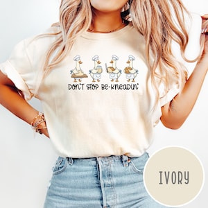 May include: Ivory-colored t-shirt featuring a whimsical illustration of four geese in chef hats, each holding baking-related items. The text "DON'T STOP BE-KNEADIN'" is printed below the geese. The shirt is a light, neutral color.