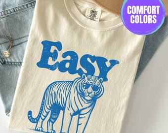 May include: A cream colored t-shirt with the text "Easy" in blue with a blue tiger wearing sunglasses. The shirt has a tag that says "Comfort Colors".