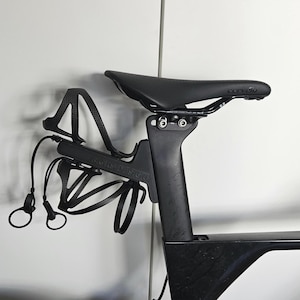 May include: A black bicycle seat mounted on a carbon fiber seat post, featuring a rear-mounted dual bottle cage system labeled MGRS DESIGN. The setup includes two black bottle cages and two black elastic cords with circular pull tabs attached.