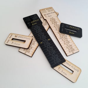 Puede incluir: Un conjunto de herramientas de prueba de corte por láser hechas de madera y acrílico negro. Las herramientas tienen medidas en milímetros y el texto "MG KERF RS TEST" y "M: ACRYLIC T: 3 mm". Las herramientas se utilizan para probar el corte por láser.
