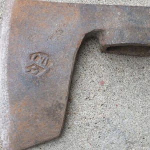 May include: Close-up of a weathered, rusty metal axe head. The axe head has a rectangular eye for the handle and a stamped emblem. The metal has a rough, textured surface and is a mix of brown and gray.