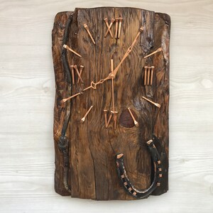 Puede incluir: Un reloj de madera rústico con marcadores de hora en números romanos hechos de clavos. La esfera del reloj está montada en una pieza de madera recuperada con una herradura unida a la parte inferior.