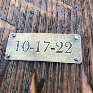 May include: A close-up of a brass plaque with the date "10-17-22" engraved on it. The plaque is attached to a wooden surface with small metal screws.