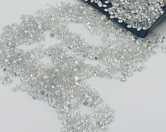 May include: A large quantity of small, clear, round diamonds are scattered on a white surface. Some of the diamonds are in a black tray.
