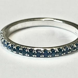 Könnte beinhalten: Ein silberner Ring mit einem Band aus kleinen, dunkelblauen Edelsteinen. Der Ring ist vor einem weißen Hintergrund platziert, der das Funkeln der Edelsteine hervorhebt. Ein zarter Schmuck.
