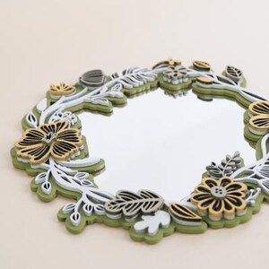 May include: A round mirror with a decorative frame made of layered wood cutouts in shades of green, white, and gold. The frame features a floral design with leaves and flowers.