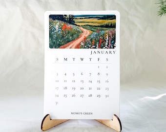 May include: A white desk calendar for January on a light wooden stand. The top features a colorful landscape painting of a dirt path through a wildflower meadow. The calendar grid displays the days of the month and the text MOMO'S GREEN at the bottom.