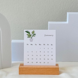 May include: A wooden desktop calendar with a white card featuring a green plant illustration and the month of January. The calendar has a simple design with the days of the week and numbers printed in black.
