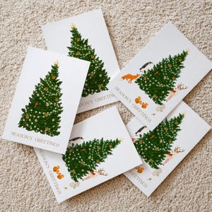Könnte beinhalten: Eine Sammlung weißer Weihnachtskarten mit einem grünen Weihnachtsbaum, geschmückt mit Lichtern und Ornamenten. Einige Karten zeigen Illustrationen von Waldtieren wie Füchsen, Hirschen und Eichhörnchen. Der Text "SEASON'S GREETINGS" ist auf jeder Karte aufgedruckt.