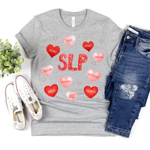May include: A light gray t-shirt with the word "SLP" in red, surrounded by red and pink hearts with speech therapy terms. The shirt is paired with blue jeans and white sneakers.
