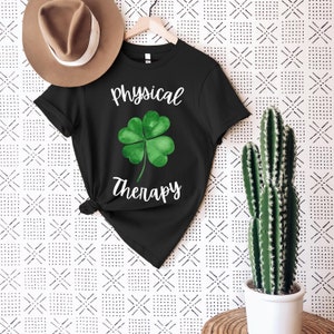 May include: Black t-shirt with the words "Physical Therapy" and a green watercolor shamrock design. The shirt is hanging on a wooden hanger. A brown hat is visible in the upper left corner.