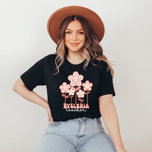 May include: Black t-shirt featuring a floral design with smiling flower faces in shades of pink and peach. The text "Dyslexia teacher" is printed below the flowers in a retro font. The person is wearing a brown hat and blue jeans.